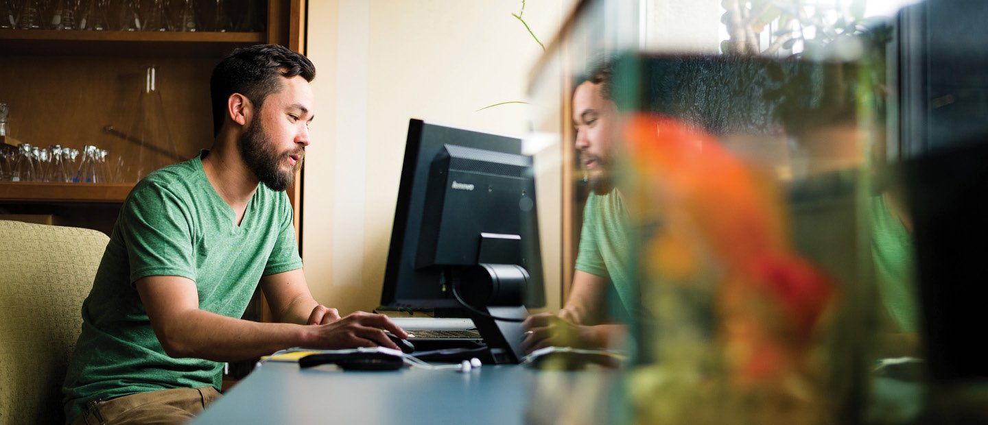 BIO-22752_GradPrograms.jpg A man seated at a computer desk with a fish tank on it.