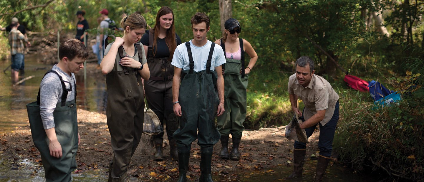 BIO-22752_bioPreserve.jpg A group of students wearing overalls and boots, observing an instructor at the Biological Preserve.