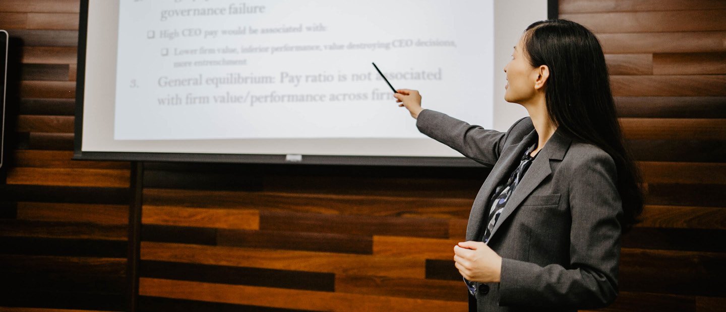 SBA-22398_Account&Finance.jpg A woman in a suit standing in front of a projector screen, pointing at a line of text on it.