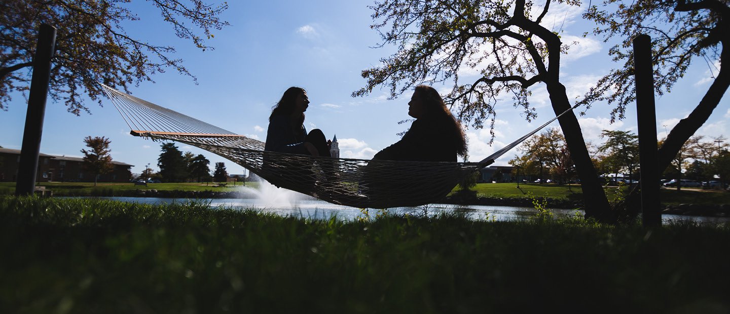 Two people seated in a hammock with a lake and fountain behind them. Two people seated, facing each other, in a hammock with a lake and fountain behind them.