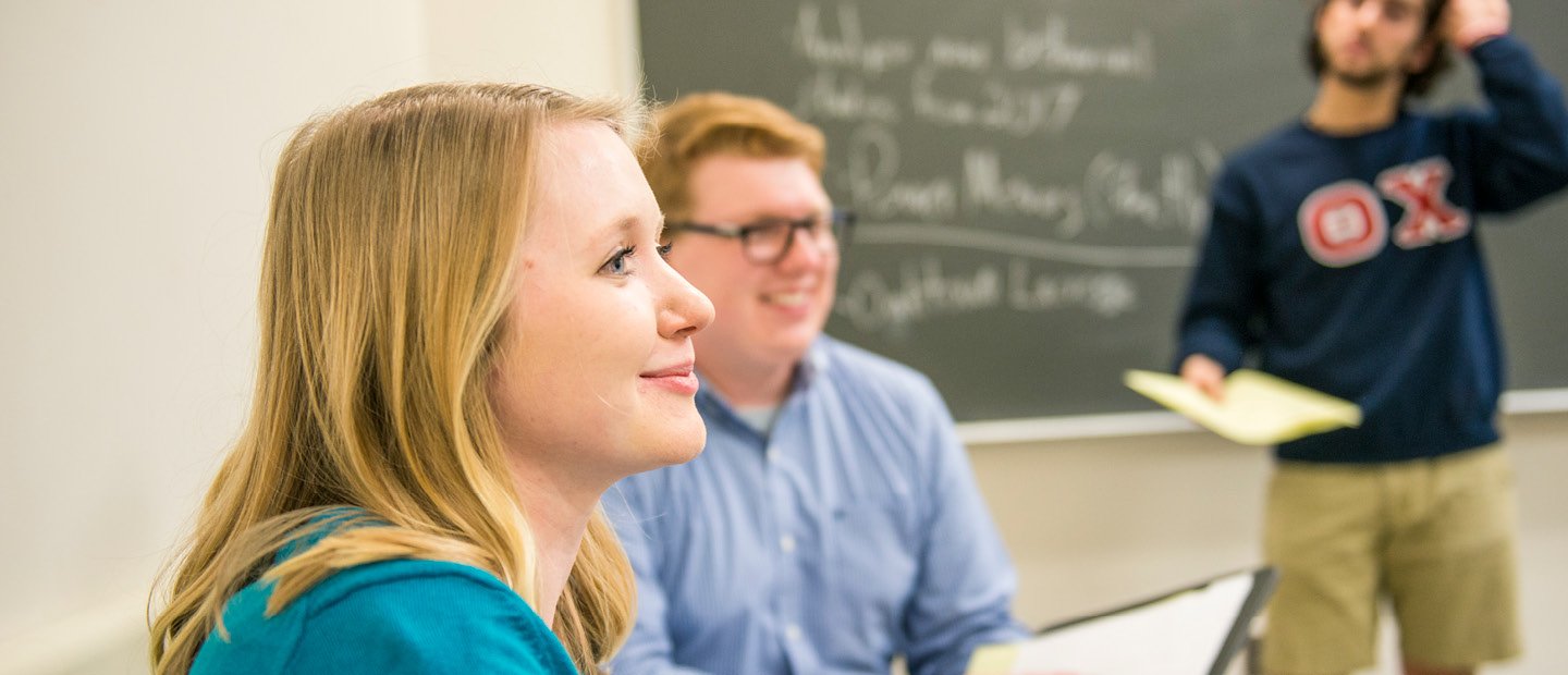 Students seated in a classroom with a professor writing on a chalkboard. A young man and woman seated, looking towards the front a classroom while a young man stands behind them by a chalkboard.