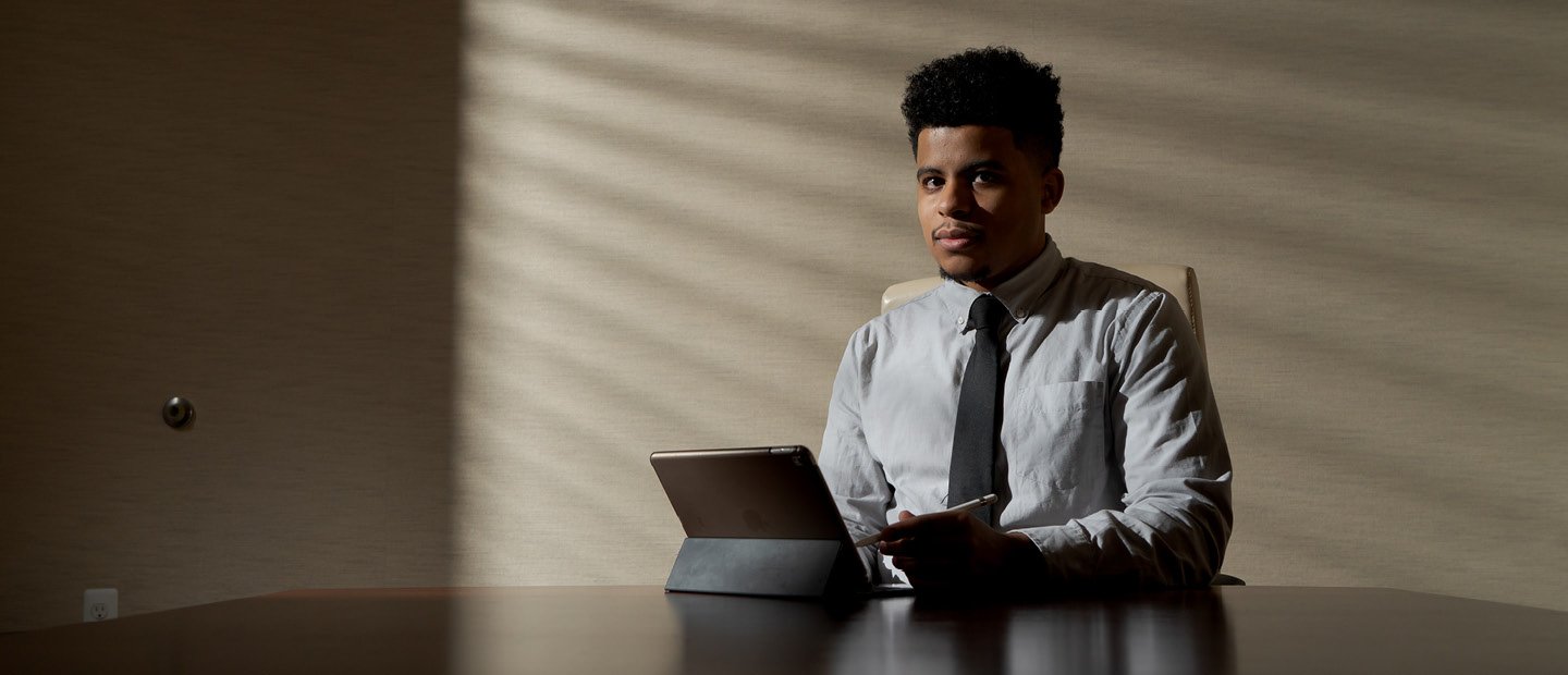 A young man seated a large table with a tablet in front of him A young man seated a large table with a tablet in front of him, looking at the camera.
