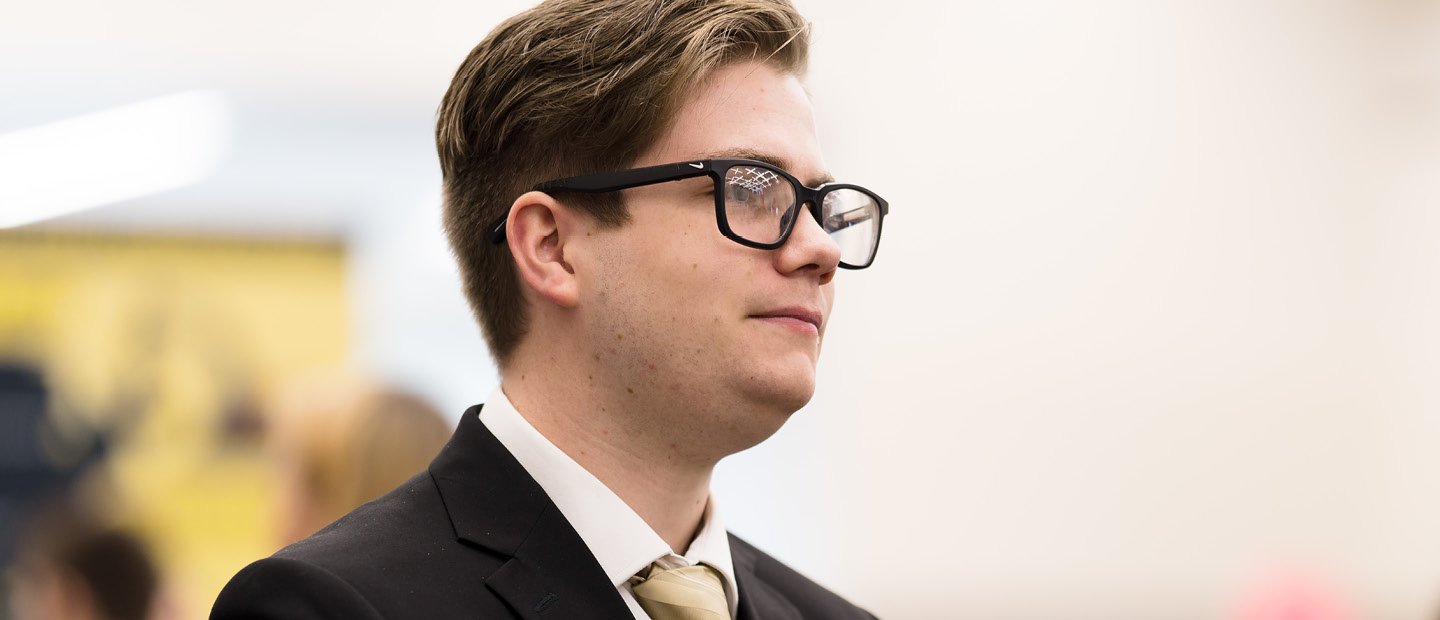 A profile photo of a man in glasses and a black suit jacket with a gold tie. A profile photo of a young man in glasses and a black suit jacket with a gold tie.