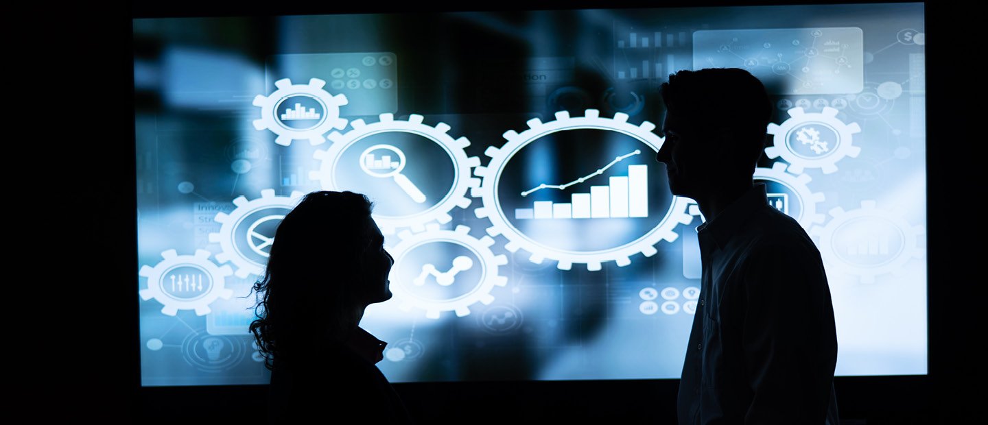 Silhouettes of two people in front of a blue screen displaying graphics. Silhouettes of two people in front of a blue screen displaying white gear and chart graphics.