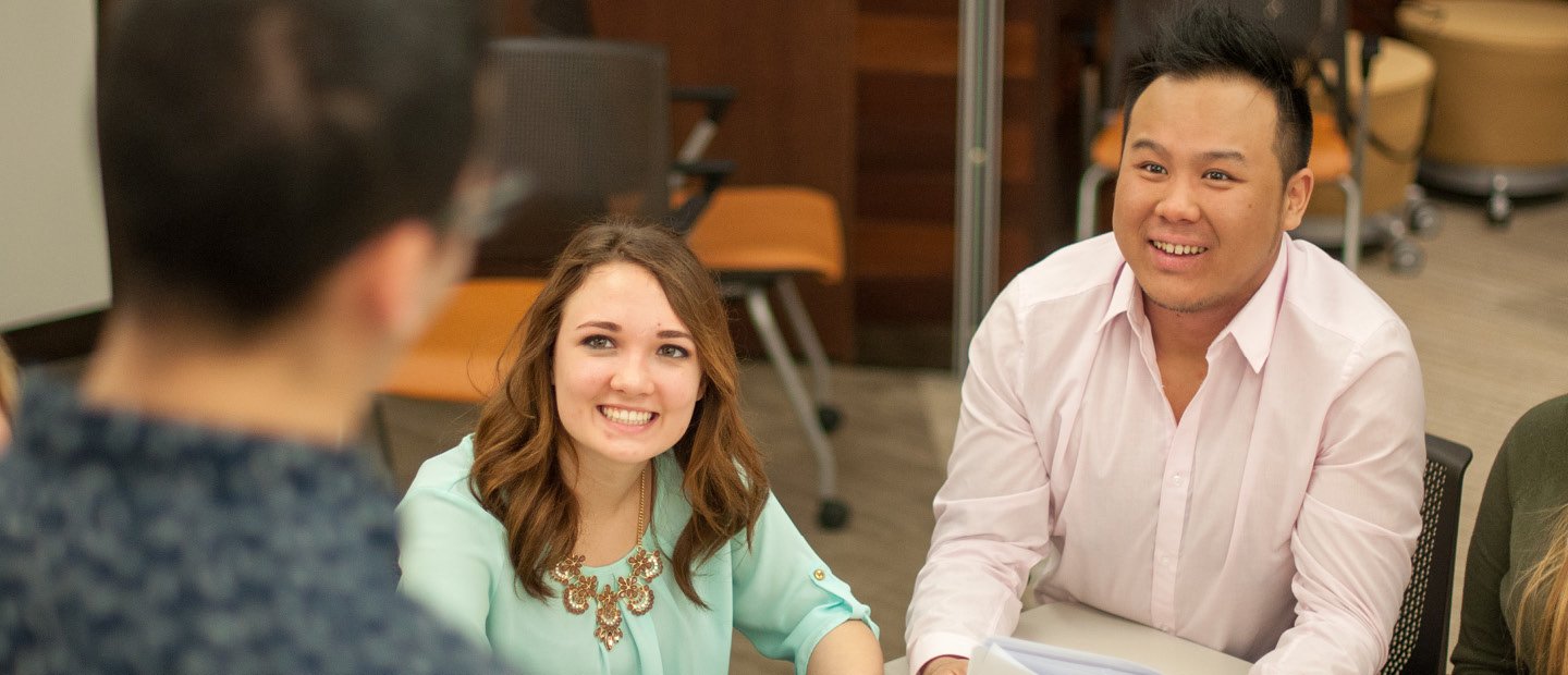 A man and woman seated at a desk, smiling at a man standing in front of them. A man and woman seated at a desk, looking up and smiling at a man standing in front of them.