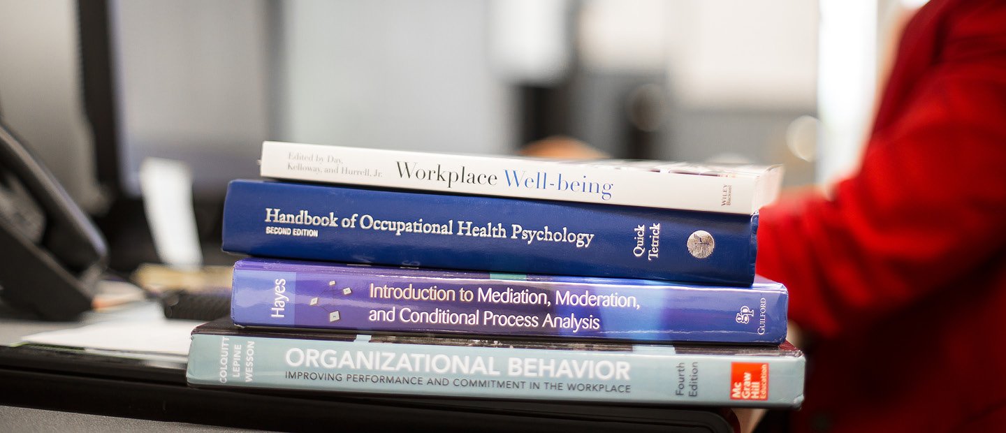 SBA-22398_Mngmnt&Market.jpg A stack of textbooks on the edge of a desk occupied by a person wearing red.