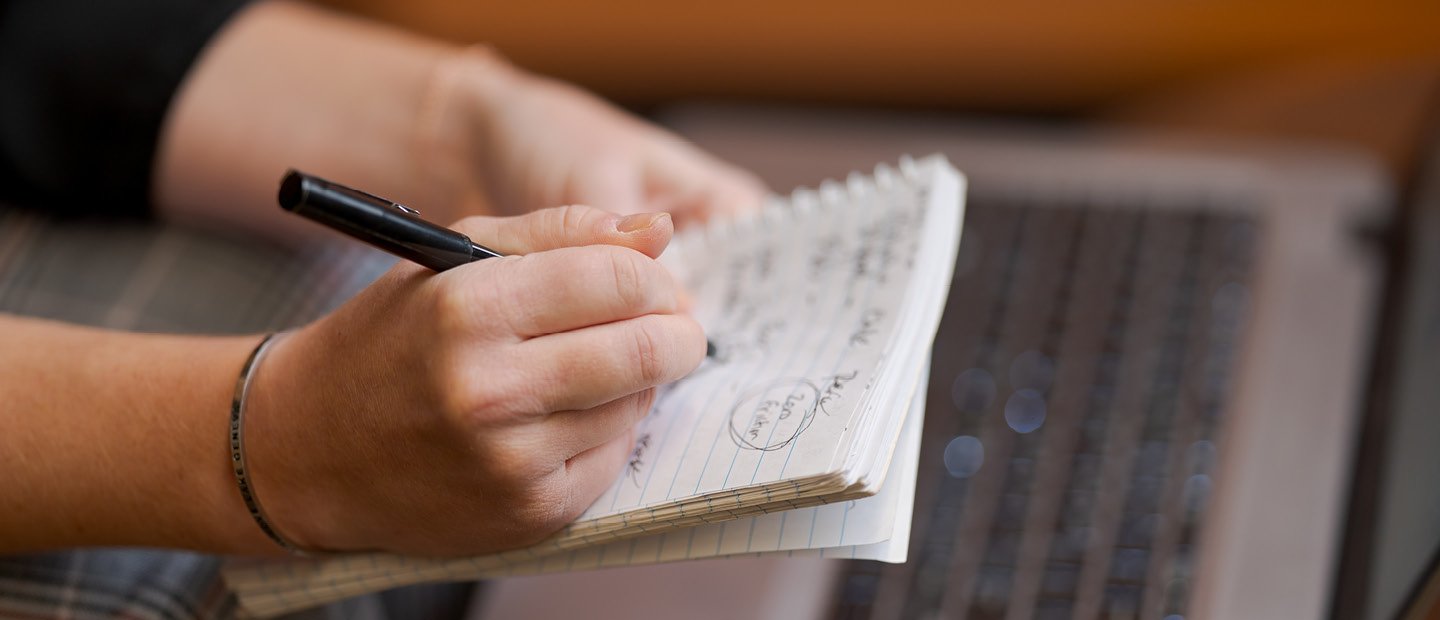 A person's hands writing on a notepad A person's hands writing on a notepad, held over an open laptop.