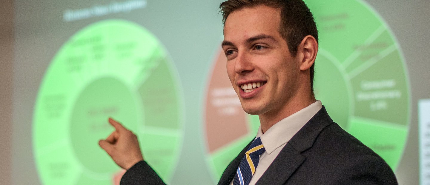 A young man in a suit, pointing to a neon green chart on a screen behind him. A young man in a suit, pointing to a neon green chart on a screen behind him.