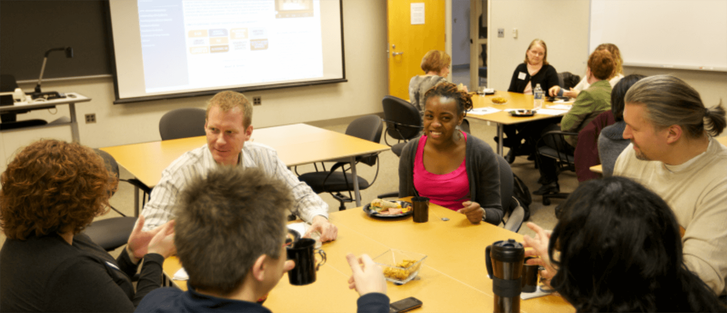 About Banner Adults seated at a two square tables in a classroom, talking and having breakfast.