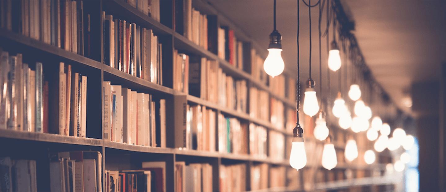 Scholarly Teachings Banner A long row of full bookshelves with hanging lights in front.