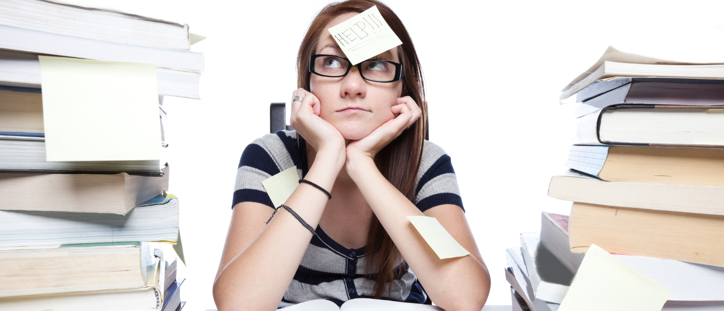 Overwhelmed woman in glasses at a desk, surrounded by high stacks of books, refl