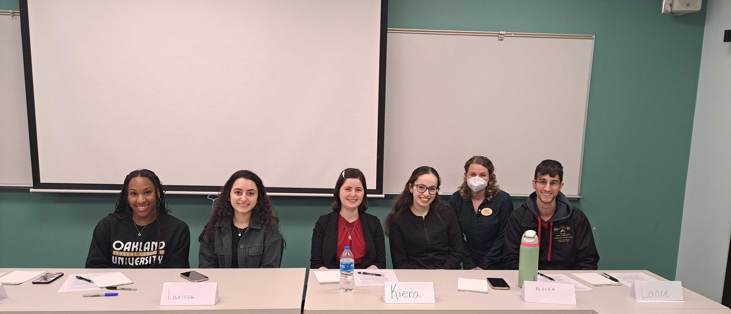 Six people sitting at a table with name tags, smiling towards the camera; a whit