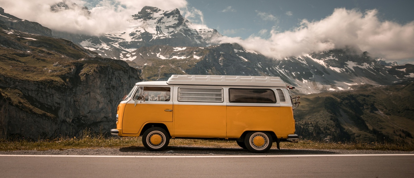 A yellow VW bus parked on the roadside with mountains in the background.