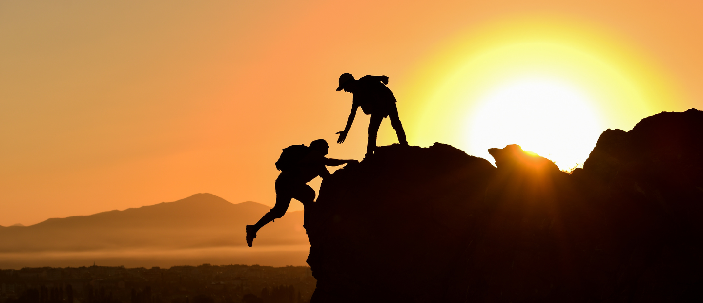 Two individuals are climbing a mountain at sunset, with one helping the other on