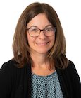 Headshot: Dark haired woman with Glasses in Dark suit Jacket and Grey top