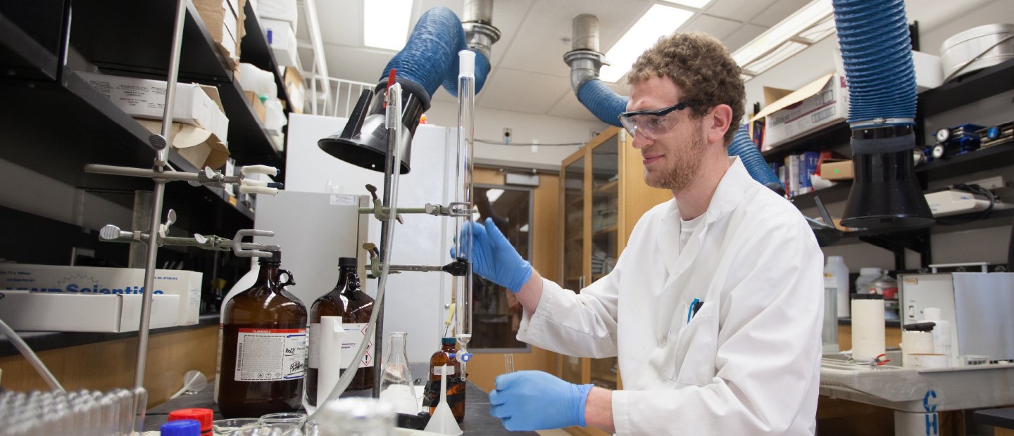 Chemistry Undergraduate Program Banner Man in goggles and lab coat works in a lab.
