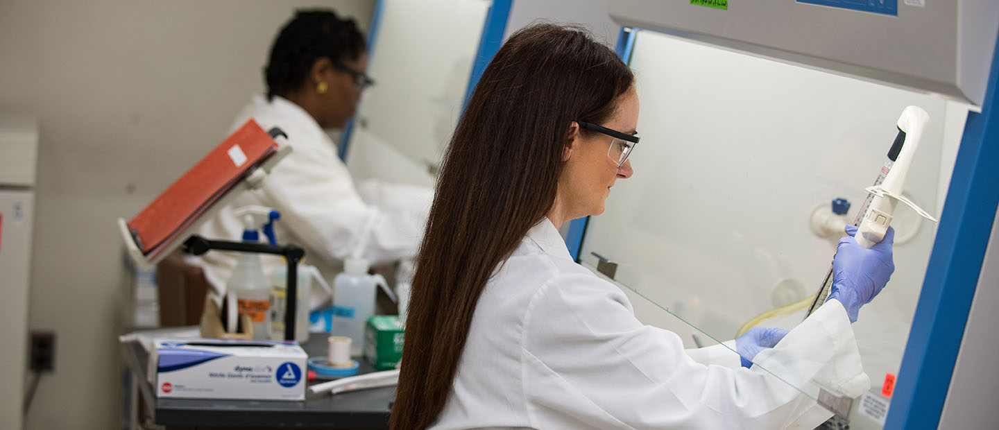 Facilities Banner two women wearing white coats, using equipment in a lab