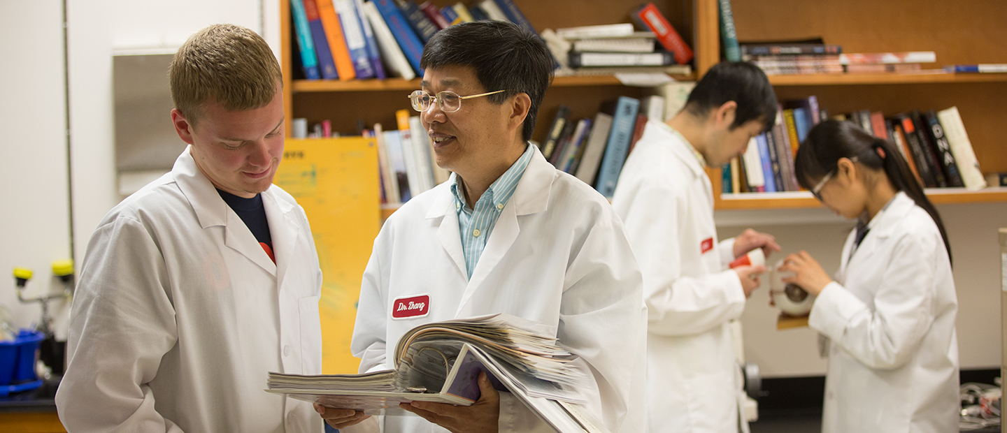 Student Research four people in white lab coats, standing in front of a book shelf, looking through a binder
