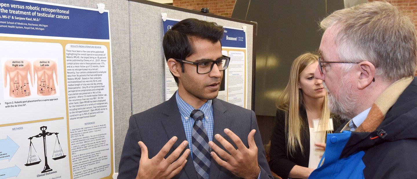 DefenseAnnouncement.jpg Student wearing a blue suit presenting his project to a professor