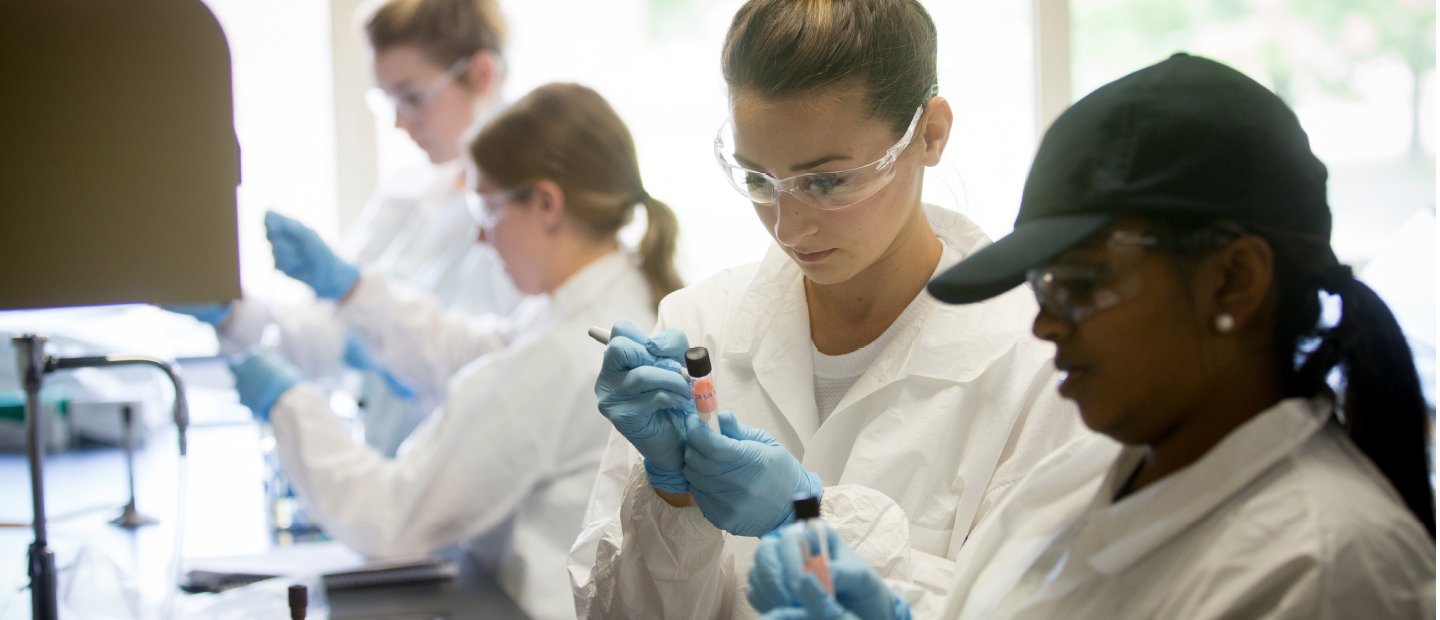 UCM-17181_Banners_doctoralstudents.jpg Students in white lab coats, safety glasses and blue gloves holding test tubes