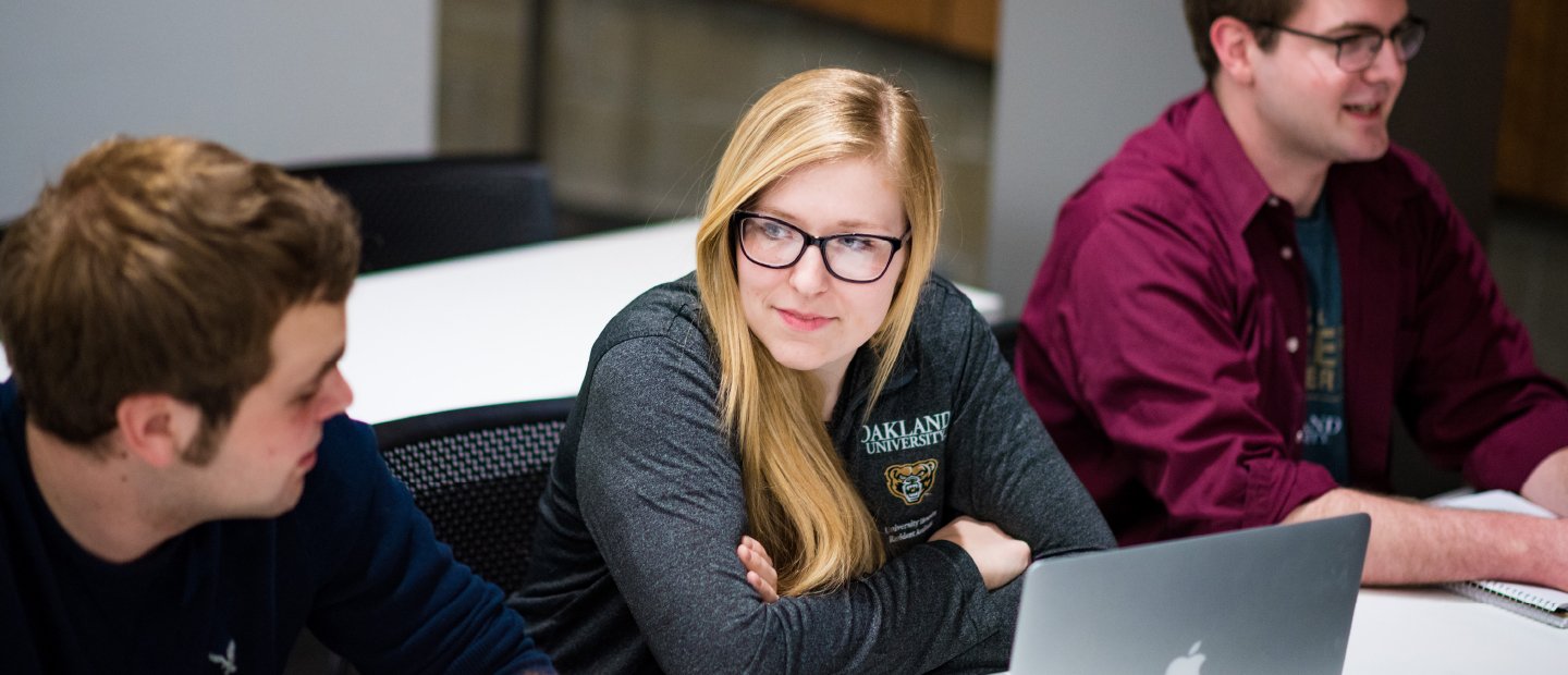 UCM-17181_Banners_nonmichigan_residencetuition_scholarship.jpg female student wearing a grey Oakland University shirt with an open laptop in front of her, male student on her right and left