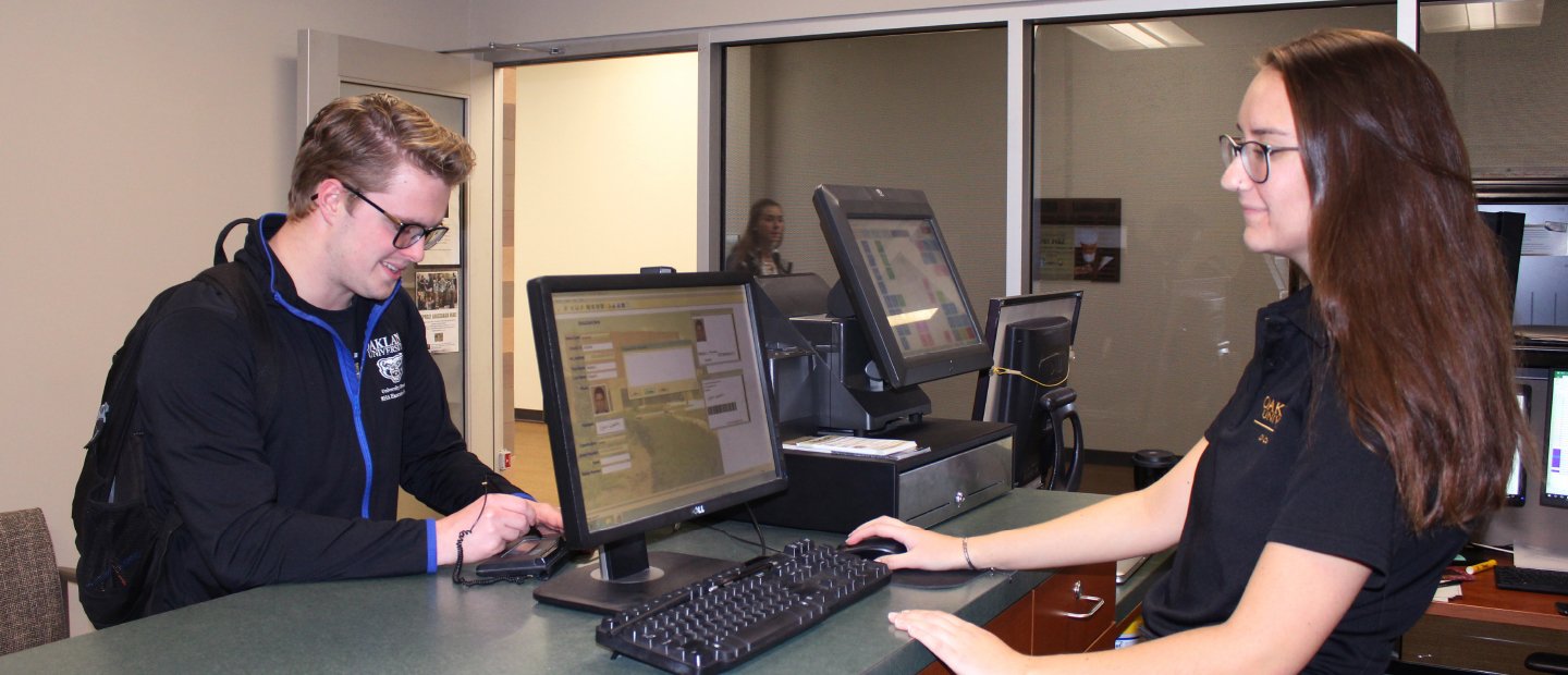 Get Your Grizzcard Banner young woman standing behind a desk with a computer, assisting a young man on the other side of the counter