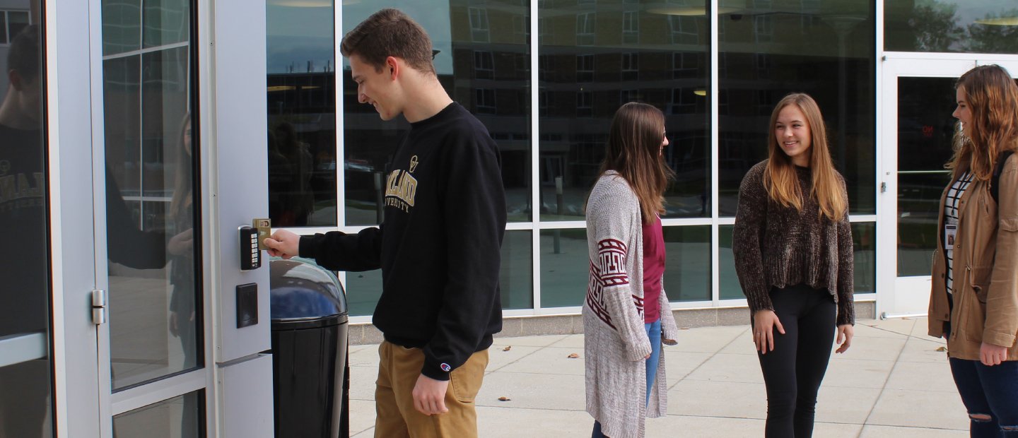 Additional Services Banner young man swiping an ID card through a reader on a wall outside, three young women in the background