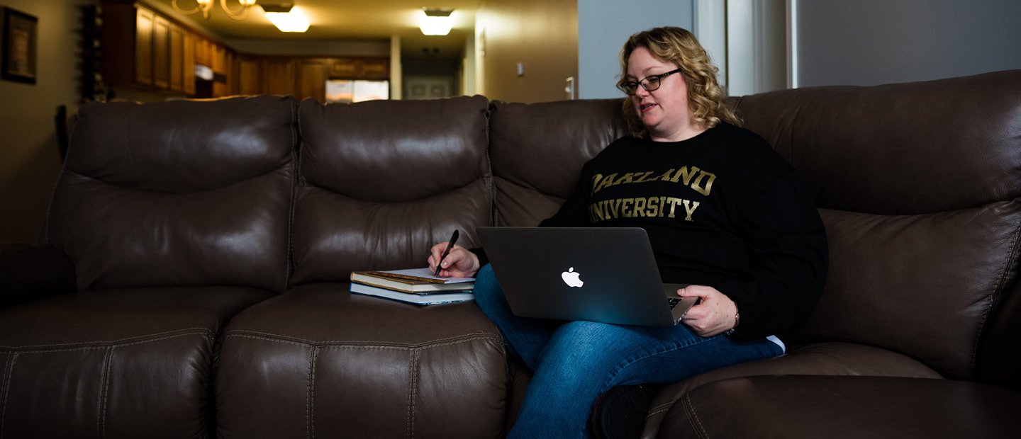 Online Student Services Banner A woman sits on a living room sofa with her laptop open.