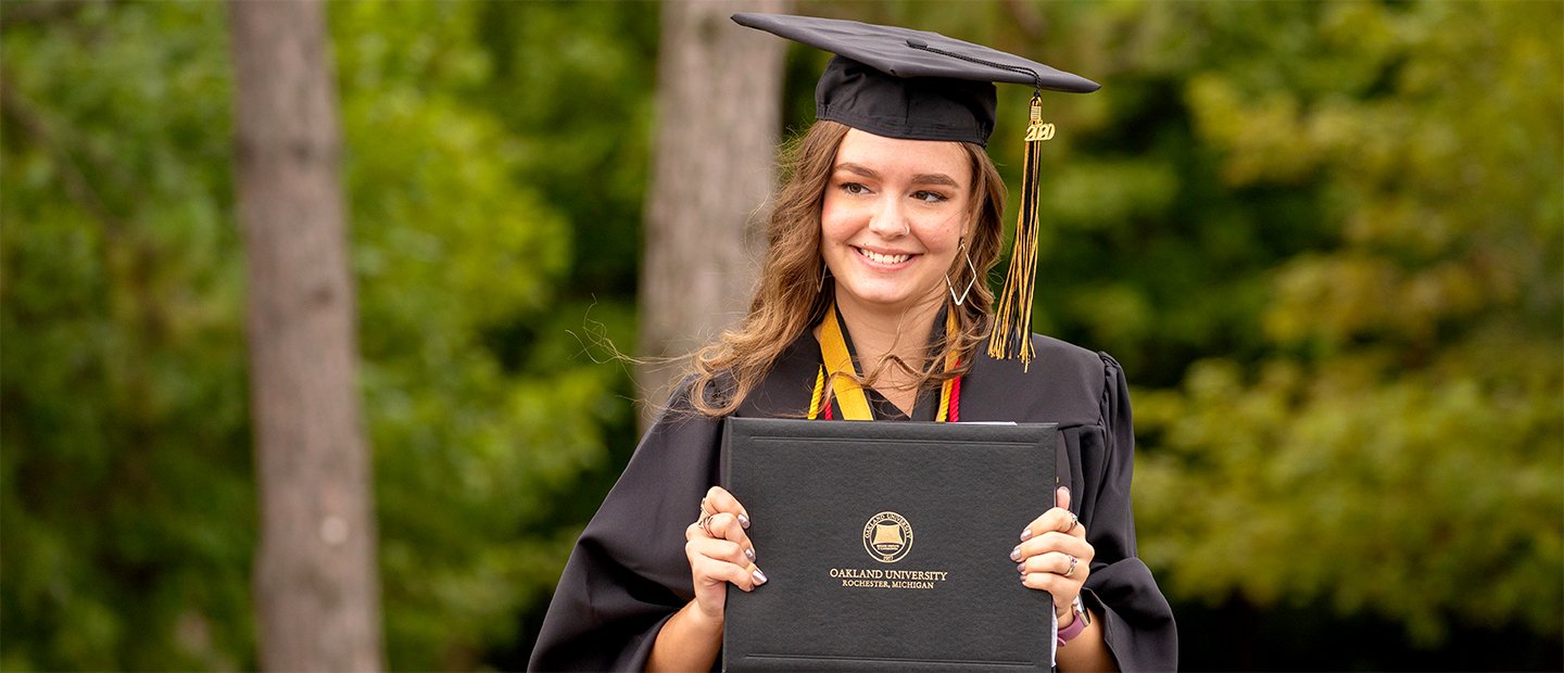 Apply for Graduation Banner Student in a cap and gown smiling at camera with diploma