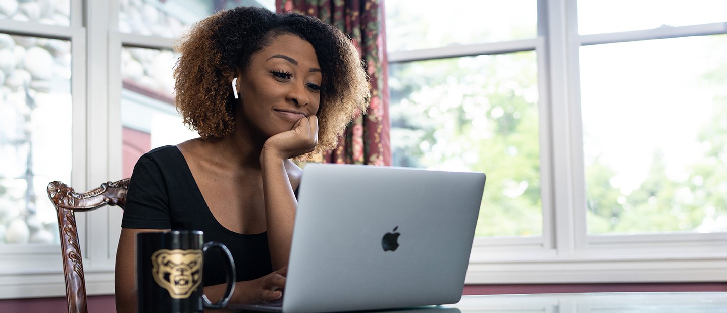 Registration Banner A female student seated at a table with an open laptop and a coffee mug with a gold Oakland University Grizz bear image on it.