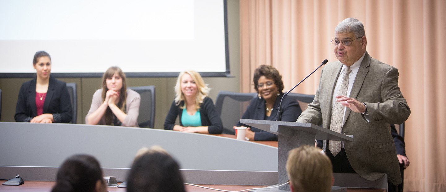 I2B Banner A man speaking into a microphone at a podium, with a panel of people seated behind him.