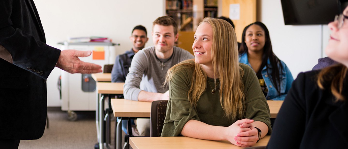 students in class classroom full of students watching a professor who is gesturing with his hands