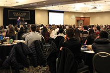 A large room full of people seated at tables, listening to a presentation.