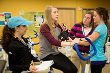 A young woman on a fitness bike with three women performing fitness evaluations on her.