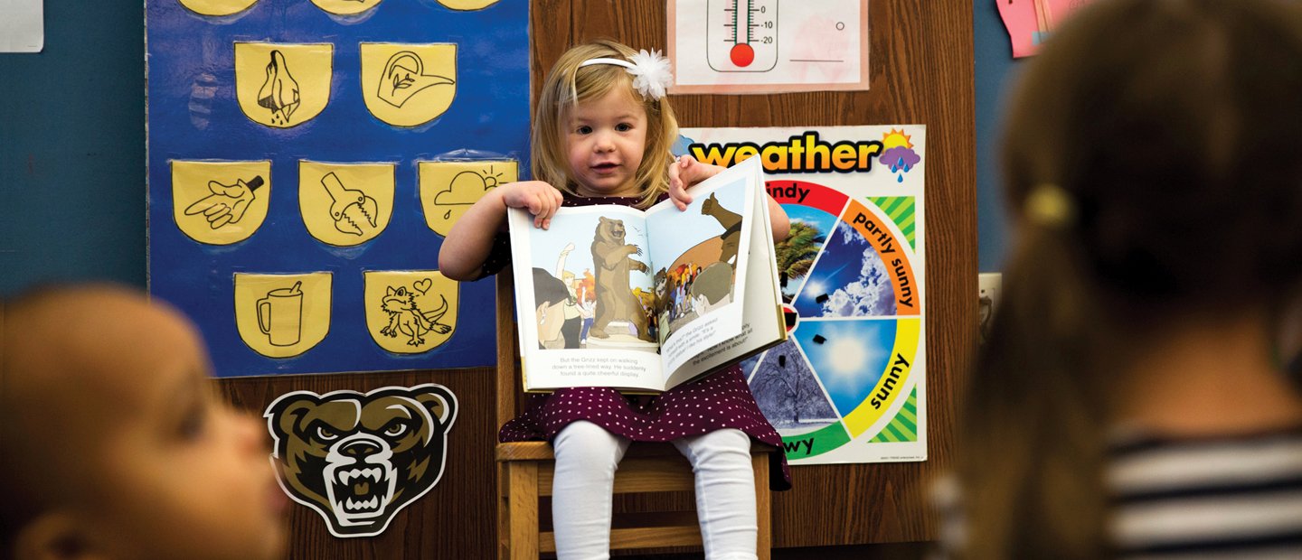 Young girl showing her classmates a book Young girl seated at the front of a classroom, showing her classmates a book.