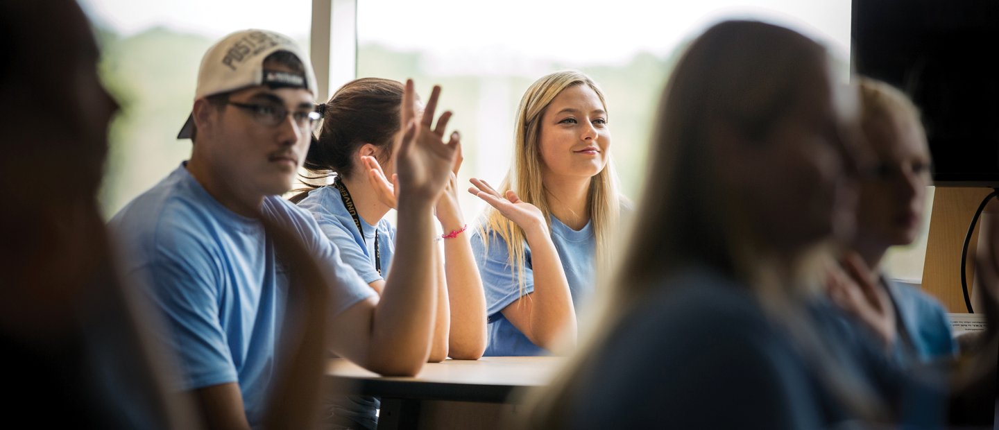 Young adults seated at desks, raising their hands Young adults in blue t-shirts, seated at desks, raising their hands.