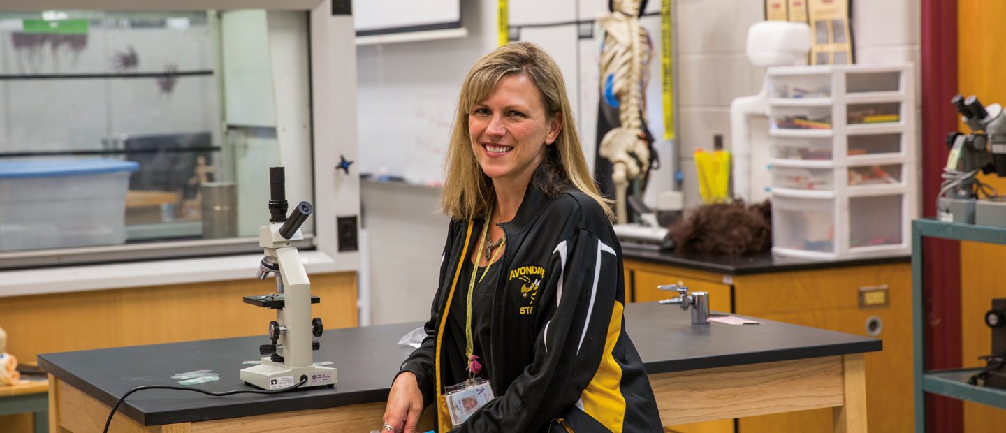 A teacher seated at a lab table with a microscope A teacher in an athletic jacket seated at a lab table with a microscope on it.