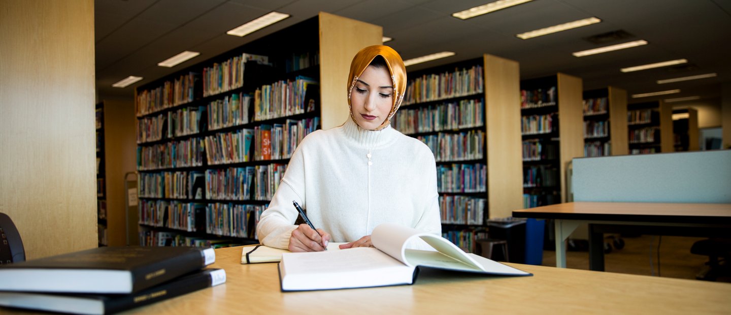 A woman seated at a table bookshelves behind her A woman seated at a table with open books, with rows of bookshelves behind her.