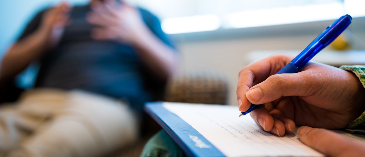 _0000_Resources and Forms.jpg A hand holding a blue pen, writing on a notepad. Another person gesturing with his hands in the background.
