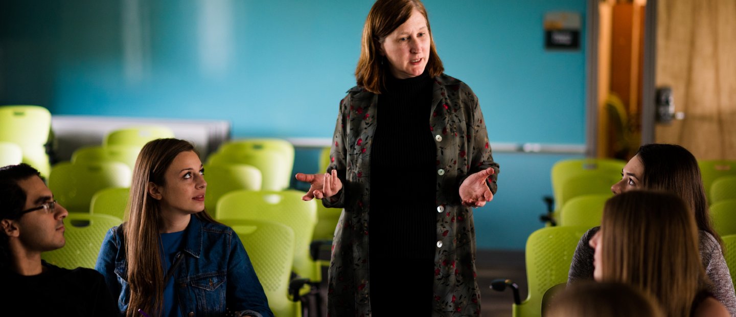 Banner - Mentors Students sitting at desks look towards a woman who stands speaking