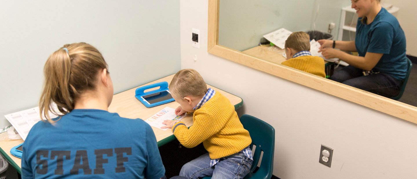 ABAClinicBanner.jpg Woman and kid sitting at desk with kid writing something on paper