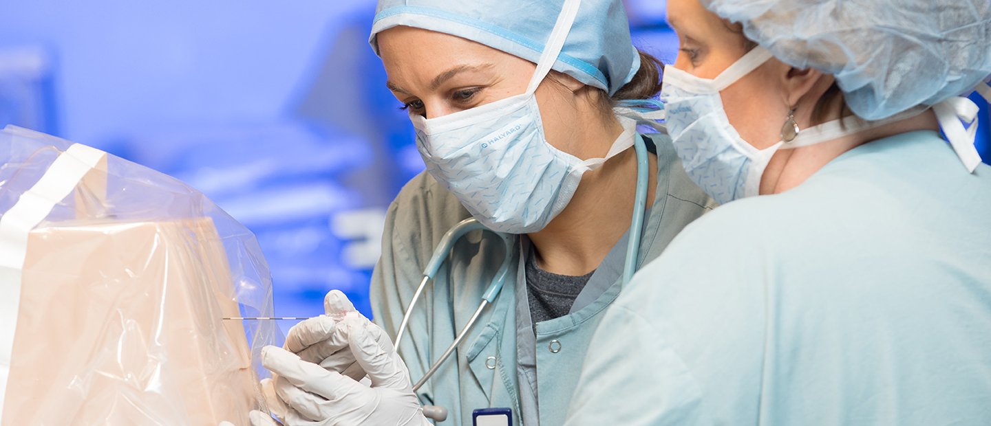 SON-19888_crna.jpg two women in surgical gowns and masks poking an object in a plastic cover