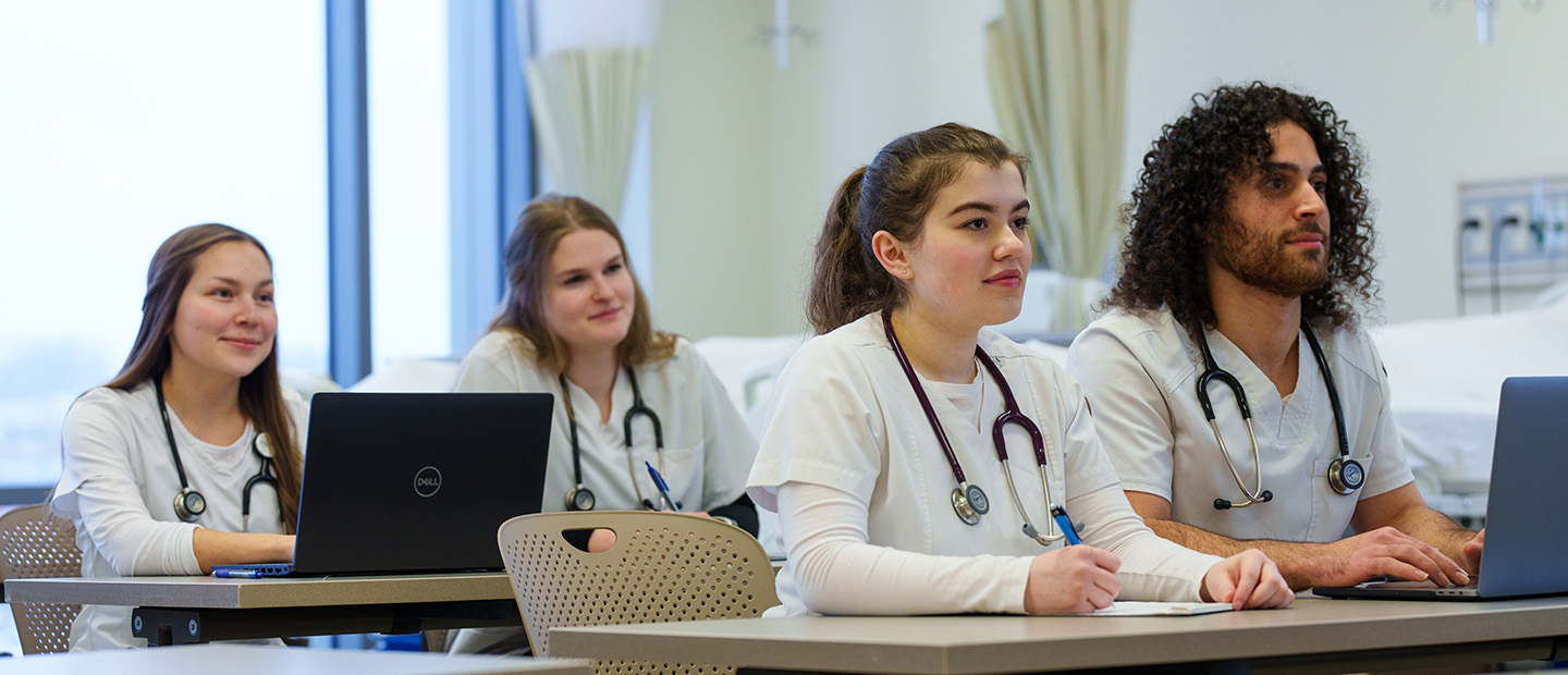 SON-25447_010 info session.jpg four students in white lab coats seated at a desk with stethoscopes